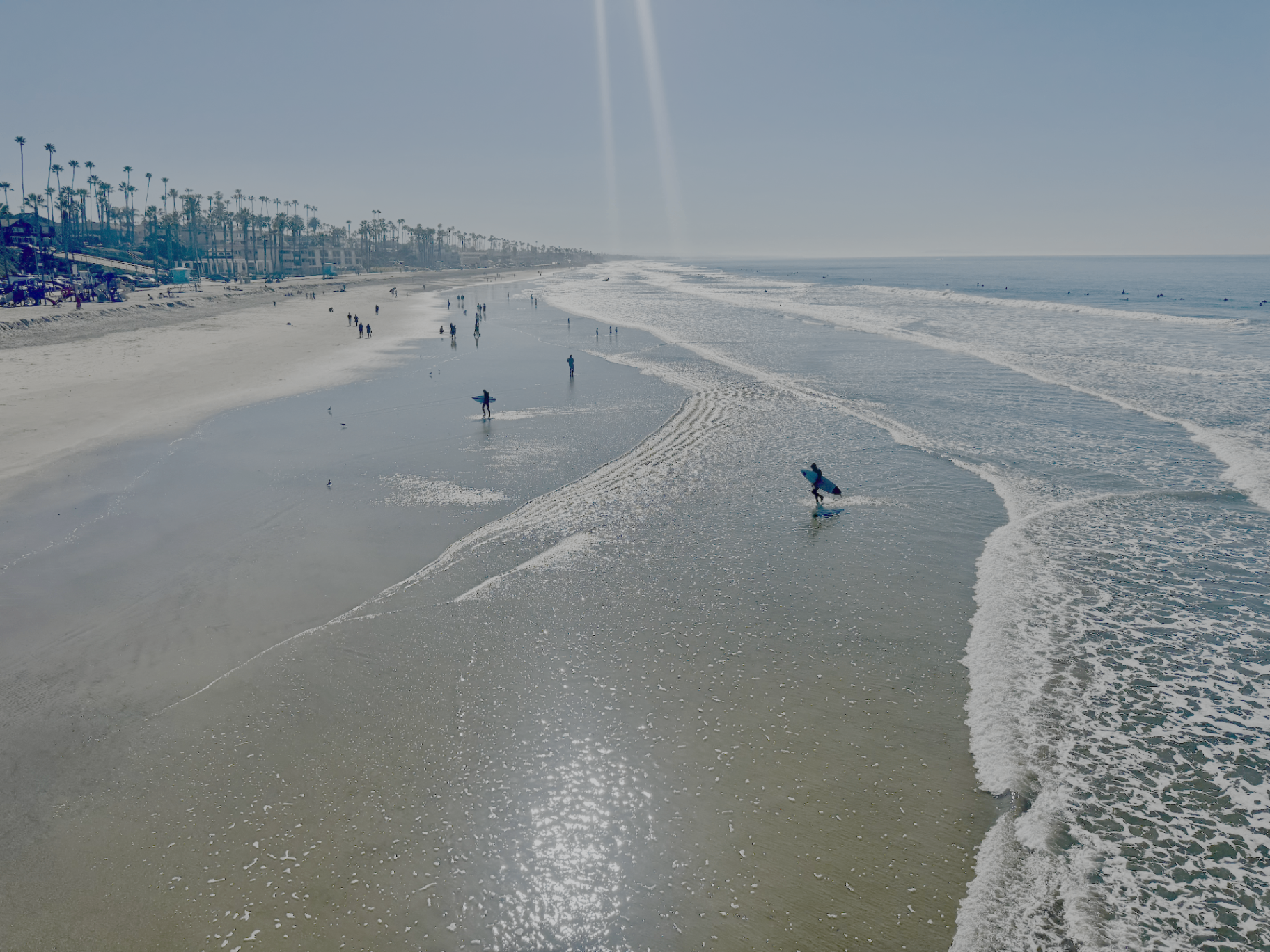 Surfers wading into the waves on a wide sandy Oceanside beach lined with palm trees